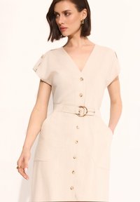 Beżowa sukienka z guzikami, z dekoltem w kształcie litery V, krótkimi rękawami, paskiem w talii oraz bocznymi kieszeniami. Posiada teksturowany materiał i mosiężne akcenty w postaci guzików.