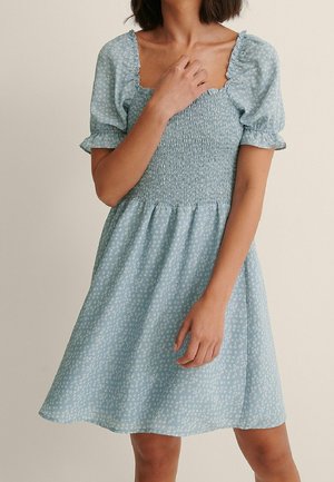 Kobieta w jasnoniebieskiej sukience z krótkim rękawem, marszczonym gorsetem i drobnym białym kwiatowym wzorem, z ręką spoczywającą blisko obojczyka i boku.