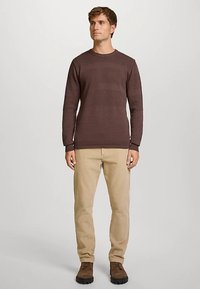 Brun strikkegenser med teksturerte striper, rund hals, og tettsittende ermer, kombinert med beige bukser og mørke sko.