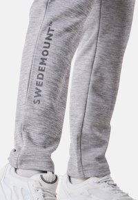 Harmaat urheiluhousut, joissa on kuvioitu kangas ja "SWEDEMOUNT" -brändipaino lahkeessa, sekä tiukat hihnat. Yhdistetty valkoisiin lenkkareihin.