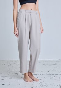 Femme portant un pantalon taille haute gris clair, ajusté, et un haut court noir, debout pieds nus sur un sol blanc texturé.