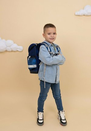 Zaino blu navy con dettagli grigi e spallacci regolabili indossato da un bambino in una camicia di denim chiaro e jeans blu, in piedi contro uno sfondo beige.