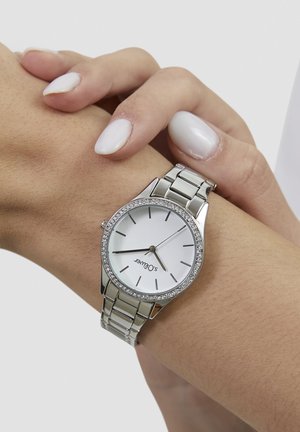 Sølvfarvet metalur med en rund hvid urskive, rhinsten-detaljer og en ledarmbånd, vist på et håndled.