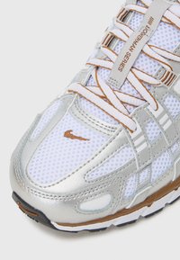 Srebrne i bijele teniske cipele s mrežastim gornjim dijelom, teksturiranim obrubom i kontrastnim vezicama. Nike logo u bakrenoj boji s strane. Gumeni potplat.