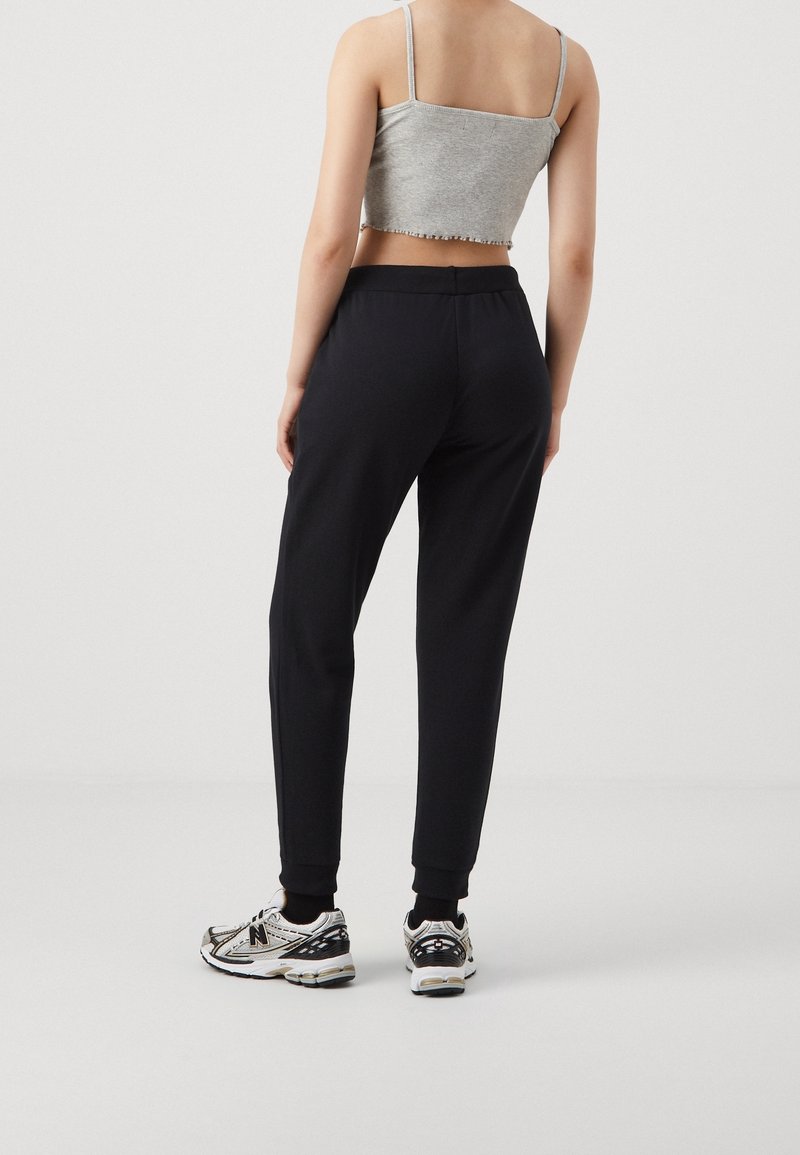 Femme portant un pantalon de jogging noir, un débardeur court gris et des baskets blanches, debout de dos sur un fond clair uni.
