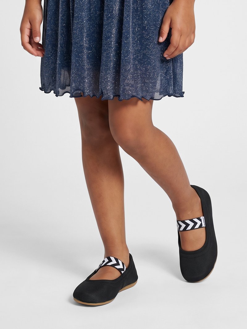 Dieťa oblečené v trblietavých modrých šatách a čiernych topánkach Mary Jane s bielymi cik-cak remienkami stojí na svetlosivej podlahe.