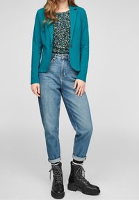 Blazer turquoise, haut à fleurs et jean taille haute bleu clair ; associé à des bottes noires à lacets de style combat. Texture lisse, coupe ajustée.