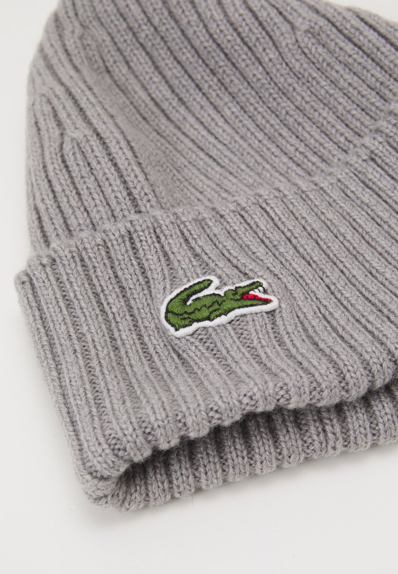 Bonnet en tricot gris avec une texture côtelée et un revers, arborant un logo de crocodile brodé vert sur le côté.