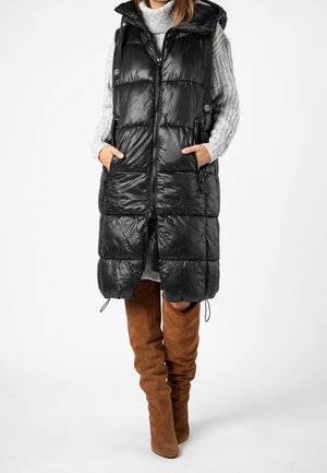 Donna che indossa un lungo gilet imbottito nero sopra un maglione dolcevita grigio con stivali marroni in camoscio alti al ginocchio, in piedi con le mani nelle tasche del gilet.