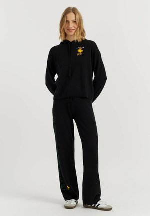 Donna che indossa una felpa nera con grafica di un uccello giallo e pantaloni neri abbinati con ricami laterali e alla caviglia, in piedi su sfondo bianco.