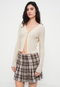 Μπεζ ριγωτό ζακάρ με μακριά μανίκια, ντεκολτέ σε σχήμα V και κυματιστό τελείωμα, σε συνδυασμό με καφέ καρό πλισέ φούστα.