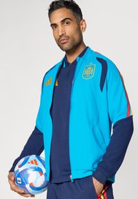 Fényes kék Adidas melegítőfelső tengerészkék díszítésekkel, nemzeti címerrel, cipzáras záródással és kontrasztos piros csíkkal az ujjakon. Mintás focilabdát tart.