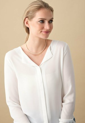 Blonde Frau in weißer Bluse trägt geschichtete Perlen- und Goldkettchen, schaut leicht nach rechts vor einem neutralen beigen Hintergrund.