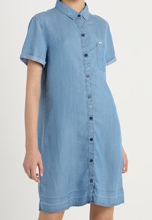 Sukienka koszulowa z denimu w jasnoniebieskim kolorze, zapinana na guziki, z krótkimi rękawami, kołnierzykiem i pojedynczą kieszonką na piersi. Rękawy podwinięte.