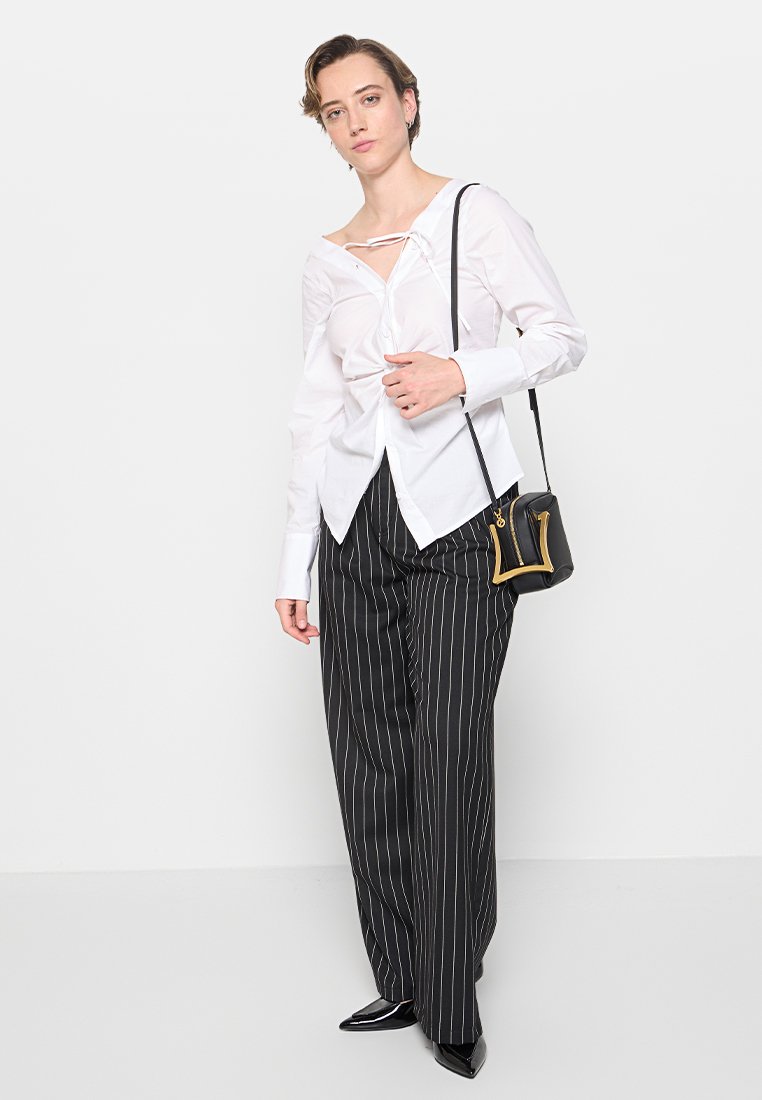 Weißes Hemd mit Frontbanddetails, lange Ärmel; schwarze Nadelstreifen-Hosen mit weitem Bein; schwarze Handtasche mit metallischen Beschlägen; schwarze spitze Schuhe.