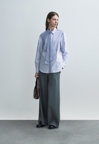 Γυναίκα όρθια που κρατά καφέ τσάντα, φοράει μπλε και λευκό ριγέ πουκάμισο με μακριά μανίκια και φαρδιά γκρι παντελόνια.