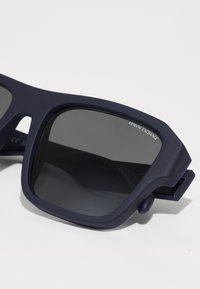 Occhiali da sole blu navy con finitura opaca, lenti rettangolari e una montatura spessa che presenta un logo in rilievo sulla parte superiore.
