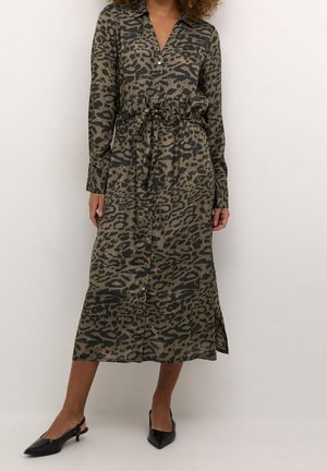 Kobieta ubrana w długą sukienkę midi z długim rękawem, zapinaną na guziki, w nadruk w panterkę, z paskiem wiązanym w talii oraz czarne, spiczaste sandały na obcasie typu slingback.