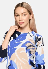 Pull en tricot avec un motif floral dans des tons de bleu, bleu marine et beige, doté d'un col rond et d'accents cloutés sur le design.