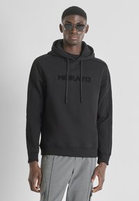 Felpa nera con logo "MORATO" ricamato, chiusura con coulisse, polsini e orlo a costine, abbinata a pantaloni grigi. Gli occhiali aggiungono contrasto.