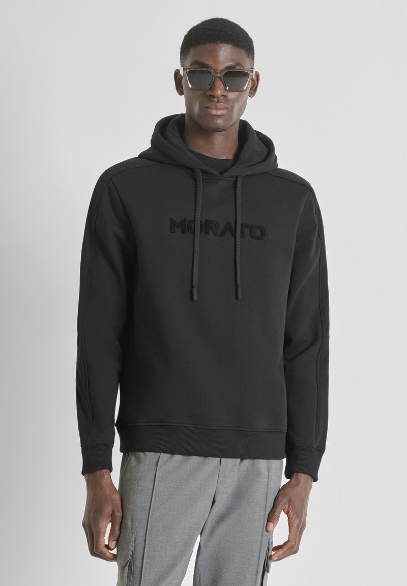 Sudadera negra con el logo bordado "MORATO", cierre con cordones, puños y dobladillo acanalados, combinada con pantalones grises. Las gafas añaden contraste.