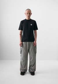 Black cotton t-shirt featuring a small white asterisk logo, paired with loose, light gray cargo pants and dark shoes.