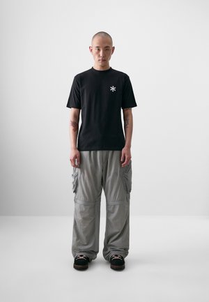 Black cotton t-shirt featuring a small white asterisk logo, paired with loose, light gray cargo pants and dark shoes.