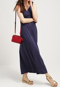 Granatowa sukienka maxi z dopasowanym gorsetem i zwiewną spódnicą. Złote sandały na koturnie z paskami. Czerwona torebka z długim paskiem.