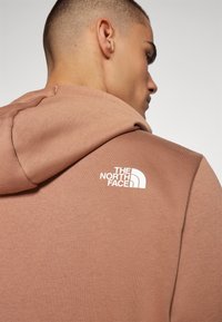 Hellbraune Kapuze mit weicher Textur, ausgestattet mit einem weißen Logo von "The North Face" auf dem oberen Rücken, Kapuzenstil mit lockerer Passform.