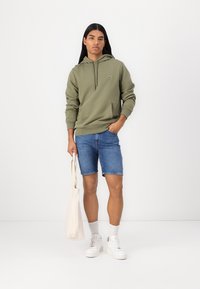 Sweatshirt à capuche vert olive avec poche kangourou, associé à un short en denim bleu, des baskets blanches et portant un tote bag crème.