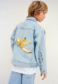 Giacca di jeans blu chiaro con un'illustrazione di una grande banana gialla sul retro. Il testo sulla manica recita: "SE SCIVOLI - RIALZATI!"