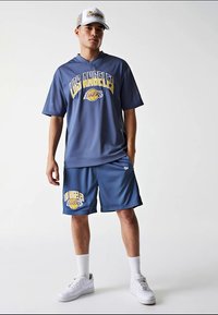 Jaunas vyras, apsirengęs mėlyna Los Andželo Lakers komandos marškinėliais, derančiais šortais, baltais sportbačiais, kojinėmis ir Lakers beisbolo kepure, stovi prieš baltą fono.