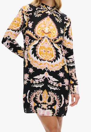 Robe noire à manches longues avec des motifs floraux et cachemire ornés en rose, jaune et blanc, portée par une femme avec une main sur la hanche.