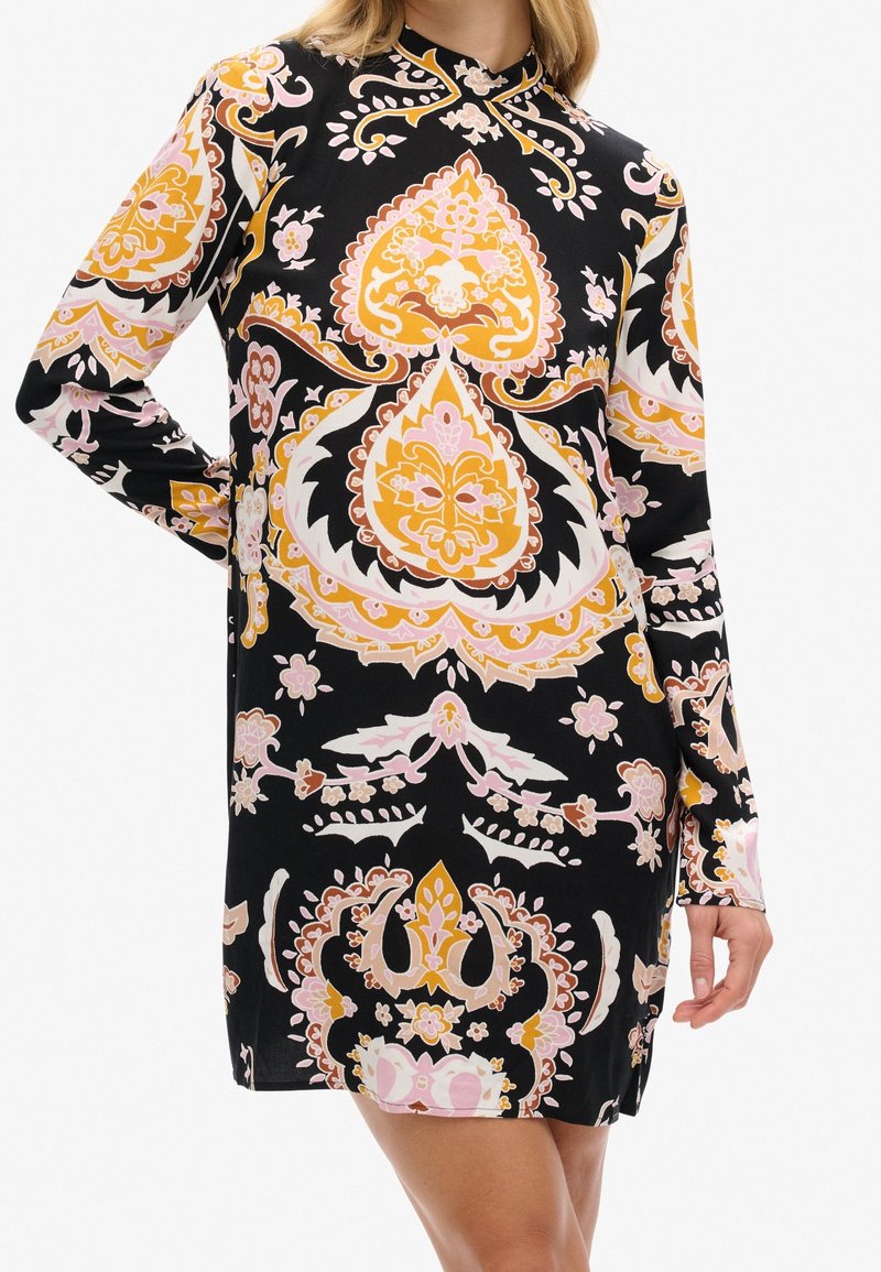 Robe noire à manches longues avec des motifs floraux et cachemire ornés en rose, jaune et blanc, portée par une femme avec une main sur la hanche.