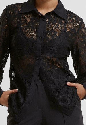 Zwarte doorschijnende kanten blouse met lange mouwen en knopen met bloemenpatroon, gedragen over een zwart topje, gecombineerd met zwarte broek, handen in de zakken.