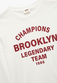Jasnoszara bluza z długim rękawem wykonana z bawełny, z wyraźnym bordowym nadrukiem: "MISTRZOWIE LEGENDARNEJ DRUŻYNY BROOKLYN 1986." Okrągły dekolt.