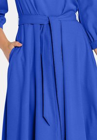 Ruka v vrecku dlhého rukávu kráľovskej modrej šiat s pásom z rovnakého materiálu zauzleným v páse.