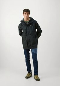 Tamsi neperšlampama striukė su kapišonu, priekinių kišenių ir logotipu. Derinama su mėlynomis kelnėmis, turinčiomis šviesesnį mėlyną panelę, ir rudos spalvos žygių batais.