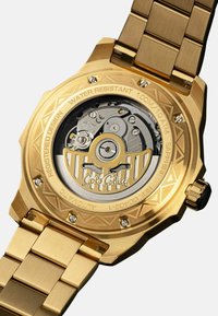 Gouden roestvrijstalen horloge met een transparante achterkant, automatische beweging zichtbaar, waterbestendig tot 100 m, ingewikkelde geometrische details.