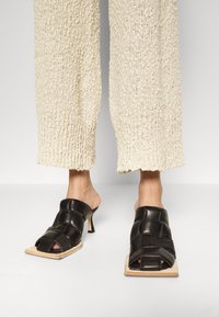 Schwarze Leder-Open-Toe-Mules mit geflochtenem Design, quadratischer Vorderseite und 5 cm Absatz. Die Holzsohle und der Knöchelschnitt unterstreichen den minimalistischen Stil.