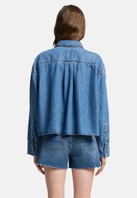 Giacca di jeans cropped di un blu chiaro con chiusura a bottoni, maniche lunghe e vestibilità rilassata. Presenta una schiena plissettata per una maggiore texture.