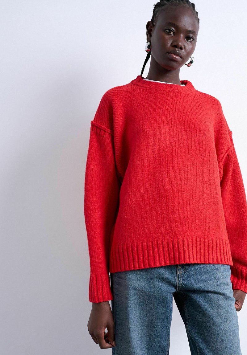 Femme portant un pull en maille rouge ample et un jean bleu sur un fond clair uni, avec des cheveux tressés et des boucles d'oreilles créoles.