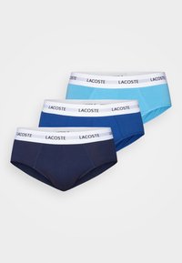 Tre par Lacoste-kalsonger för män i marinblått, mörkblått och ljusblått. Varje par har en vit elastisk midjeband med Lacoste-logotypen.