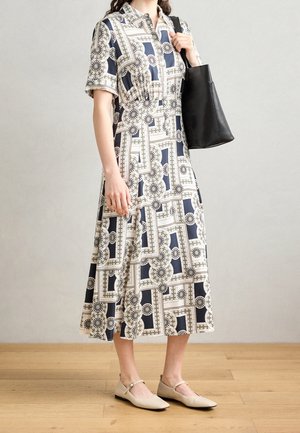 Vrouw draagt een midi-jurk met patroon en beige platte schoenen, houdt een zwarte tas over haar schouder, staand op een houten vloer.