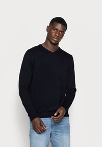 Suéter negro de pico hecho de tejido de punto, con mangas largas, puños y dobladillo acanalados. Lleva puesto sobre unos jeans azul claro.