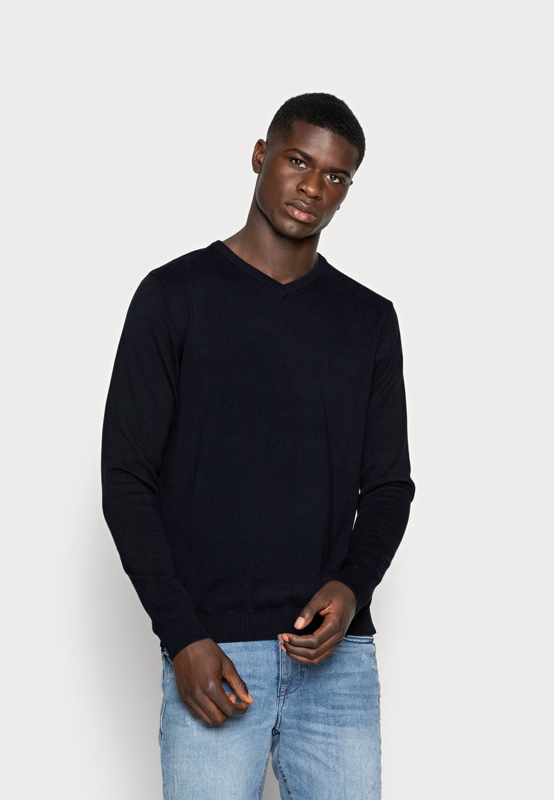 Suéter negro de pico hecho de tejido de punto, con mangas largas, puños y dobladillo acanalados. Lleva puesto sobre unos jeans azul claro.