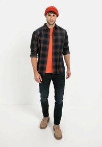 Rutede flannel skjorte i sort og rød, orange t-shirt underneath, mørke jeans og tan støvler. Strikket rød hue på hovedet. Frontvinkel.