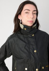 Jachetă impermeabilă neagră, cu guler înalt, accesorii aurii, nasturi frontali, două buzunare frontale și un detaliu contrastant la interiorul gulerului.