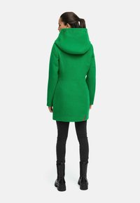 Green fleece coat with a high collar and hood, side seams, and a small logo detail at the back. Paired with black skinny jeans and boots.