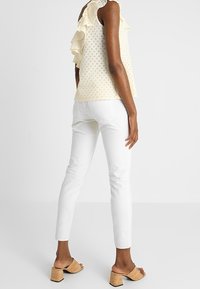 Kvinna som bär en ärmlös krämfärgad top med ögonhål och volanger samt vita slim-fit byxor, stående i bruna klackade sandaler med öppen tå på en enkel bakgrund.